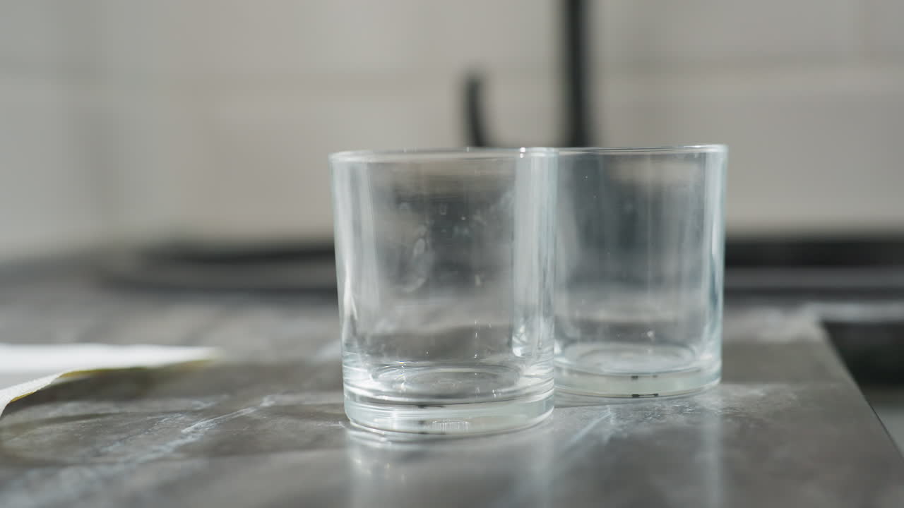 Close up of person gently dropping transparent glass cups on marble counter top in bright kitchen with soft blur background and elegant manicured fingers