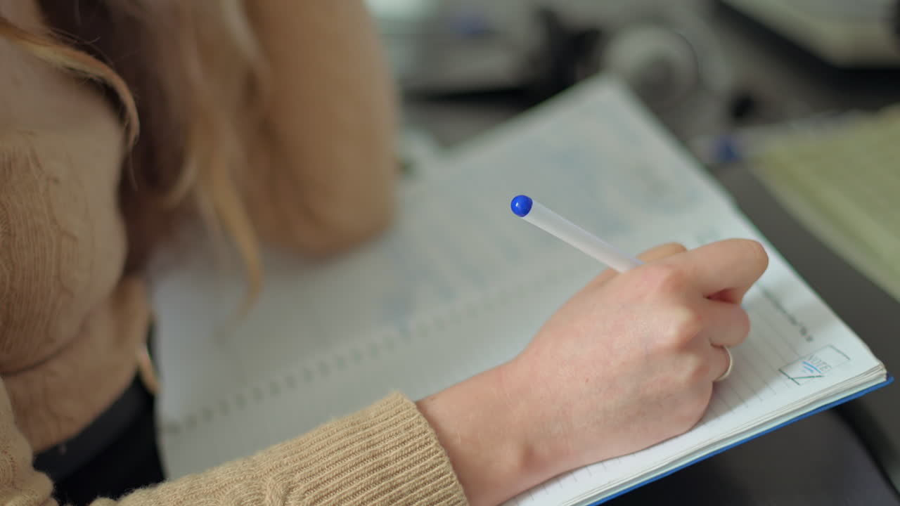 A person is focused on taking notes in a notebook at a desk. The background shows a study area filled with various supplies and materials, creating a productive atmosphere
