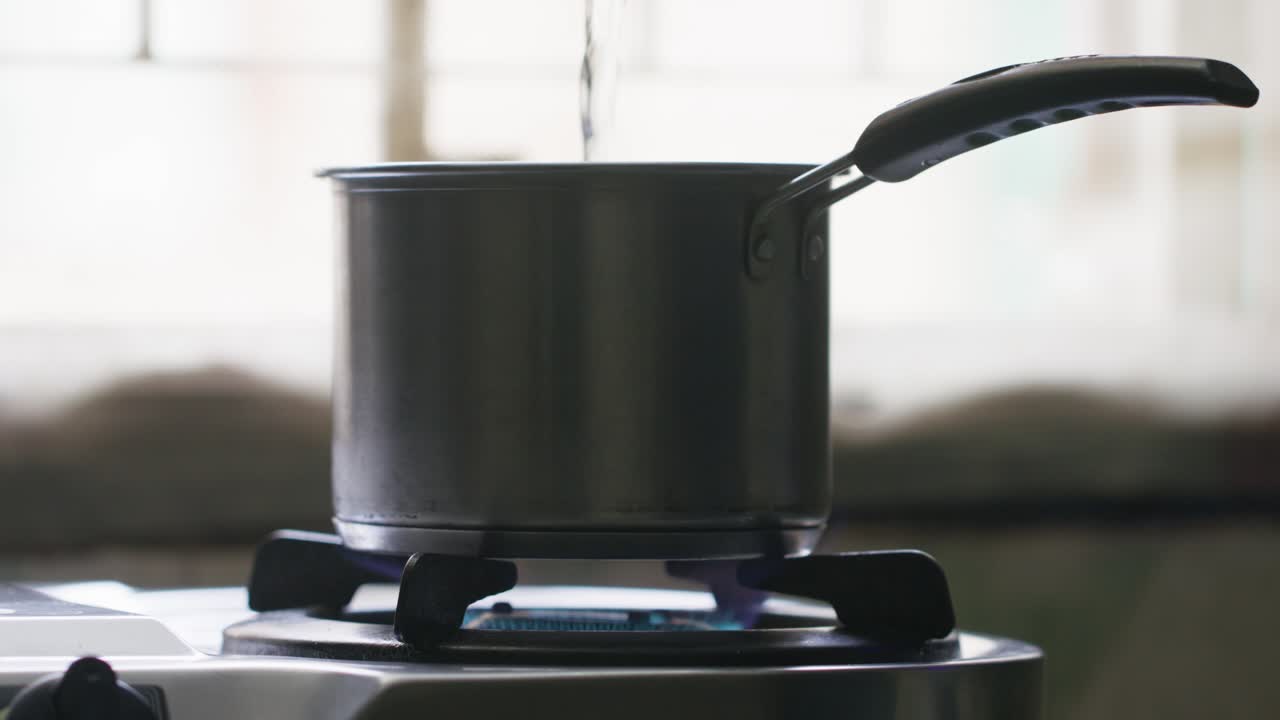 A close-up of a hand pouring water into a metal cooking pot on the lit stove against a blurred background with natural light