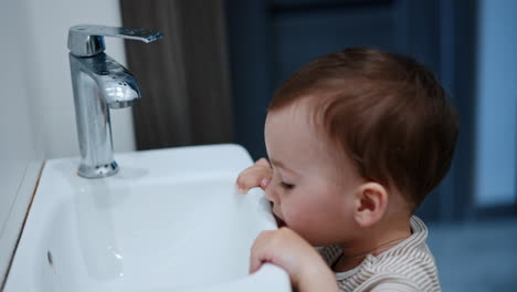 cute brown-haired toddler boy stands holding by the sink. little baby plays with water and tap. close up.