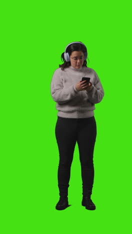 vertical video full length shot of woman wearing wireless headphones streaming music from mobile phone against green screen