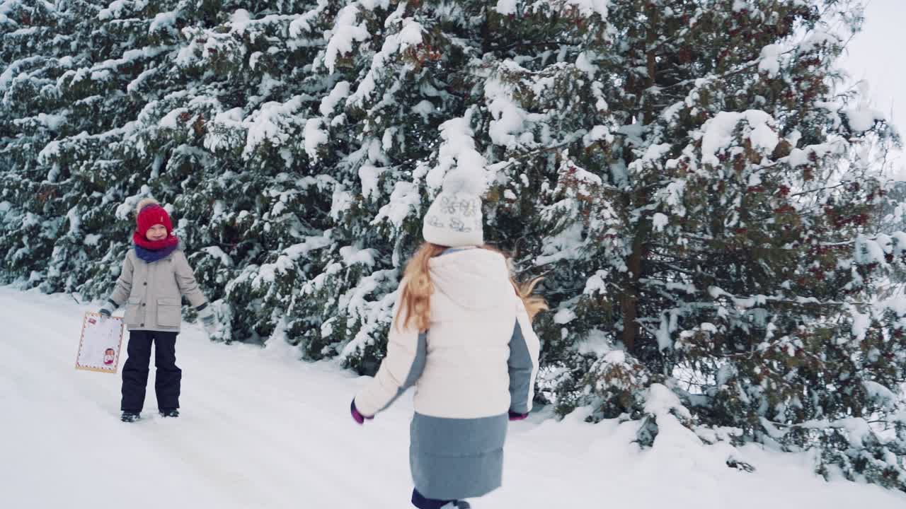 A little boy with an envelope in his hands is waiting for a girl who runs to him on the background of a winter forest on the street. And children are running along a snowy path together on the background of tall fir-trees