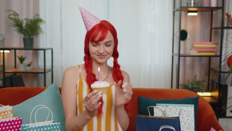 happy redhead girl celebrating birthday anniversary party, blowing candle on small cake cupcake home