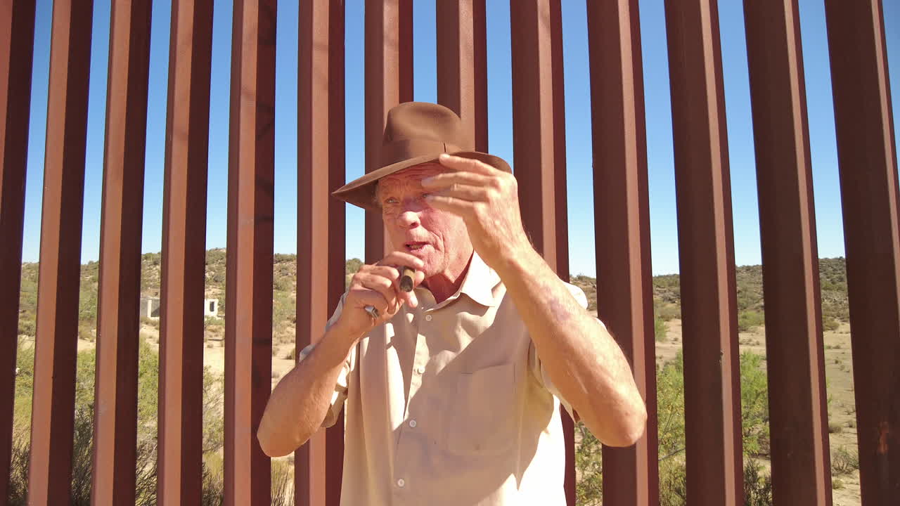 Older Caucasian man puts on fedora hat and smokes cigar in front of border fence separating USA and Mexico in Jacumba, California.