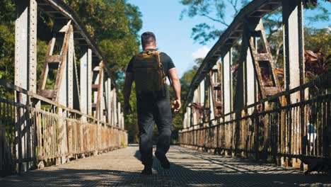 Young-man-traveler-with-backpack-walking-on-rusty-old-bridge-outdoors-on-sunny-summer-day