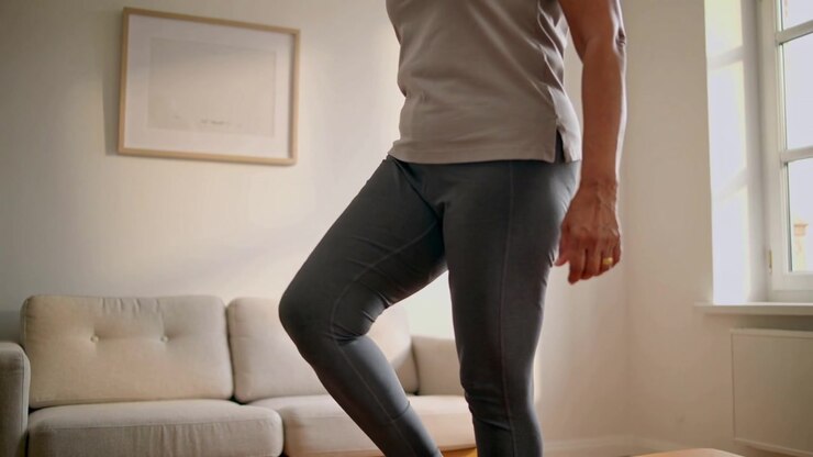 Starting senior lifting knee, balancing on foot in living room, wearing leggings, ring, for balance