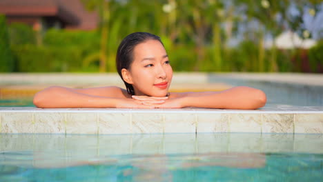 A-close-up-of-a-pretty-young-woman-with-her-head-resting-on-her-arms-along-the-edge-of-a-swimming-pool-raises-her-head-and-opens-her-eyes