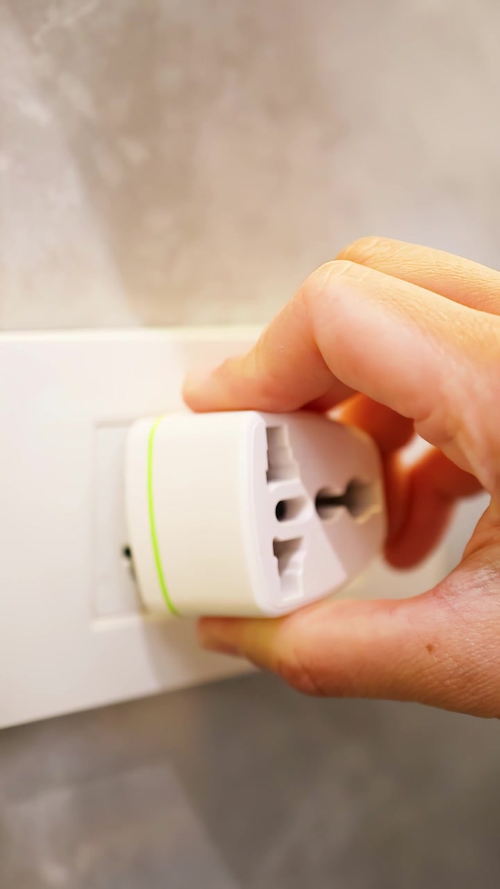 A hand inserts a white universal travel adapter into a wall outlet with soft lighting