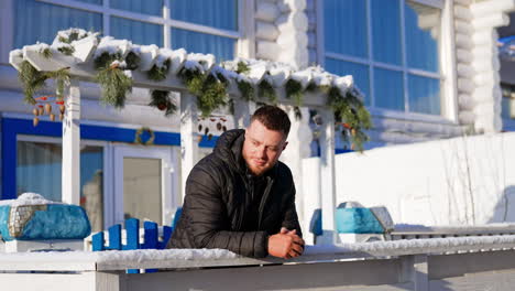 bearded caucasian man wearing black jacket stands outdoors leaning on the fence. man enjoys nice sunny day in winter.