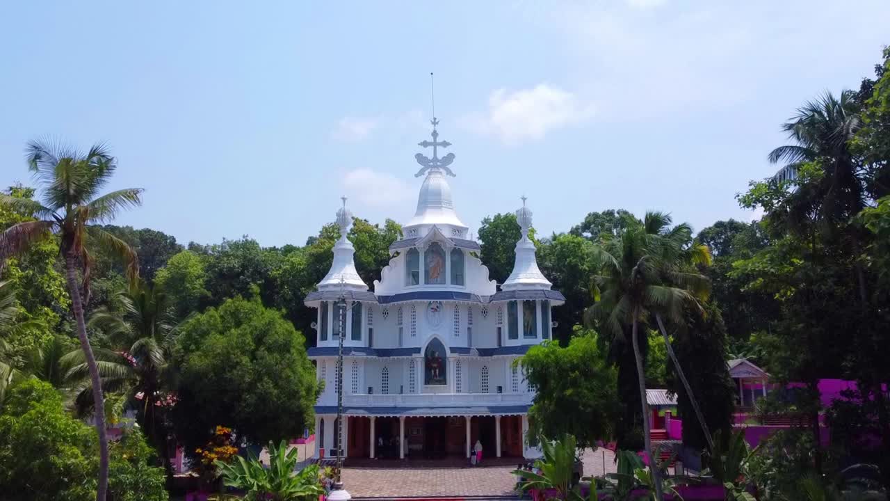 Majestic aerial footage tilting up a beautiful, ornate white church in Kerala. Its unique Syro-Malabar architecture stands out against lush tropical greenery and a clear blue sky, creating a spiritual