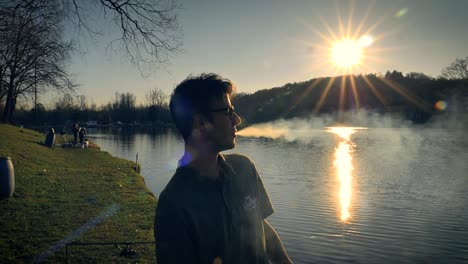 jonge man met een bril die bij zonsondergang naast een meer staat en een sigaret rookt, ademt uit terwijl hij een stroom rook uit zijn mond blaast