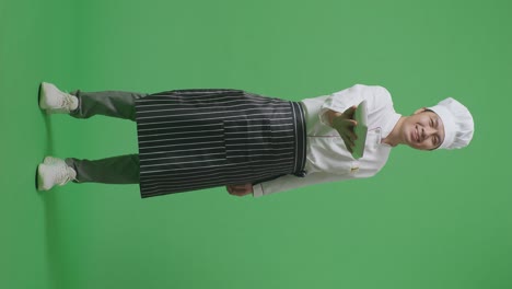 full body of asian man chef smiling and giving a dish to camera while standing in the green screen background studio