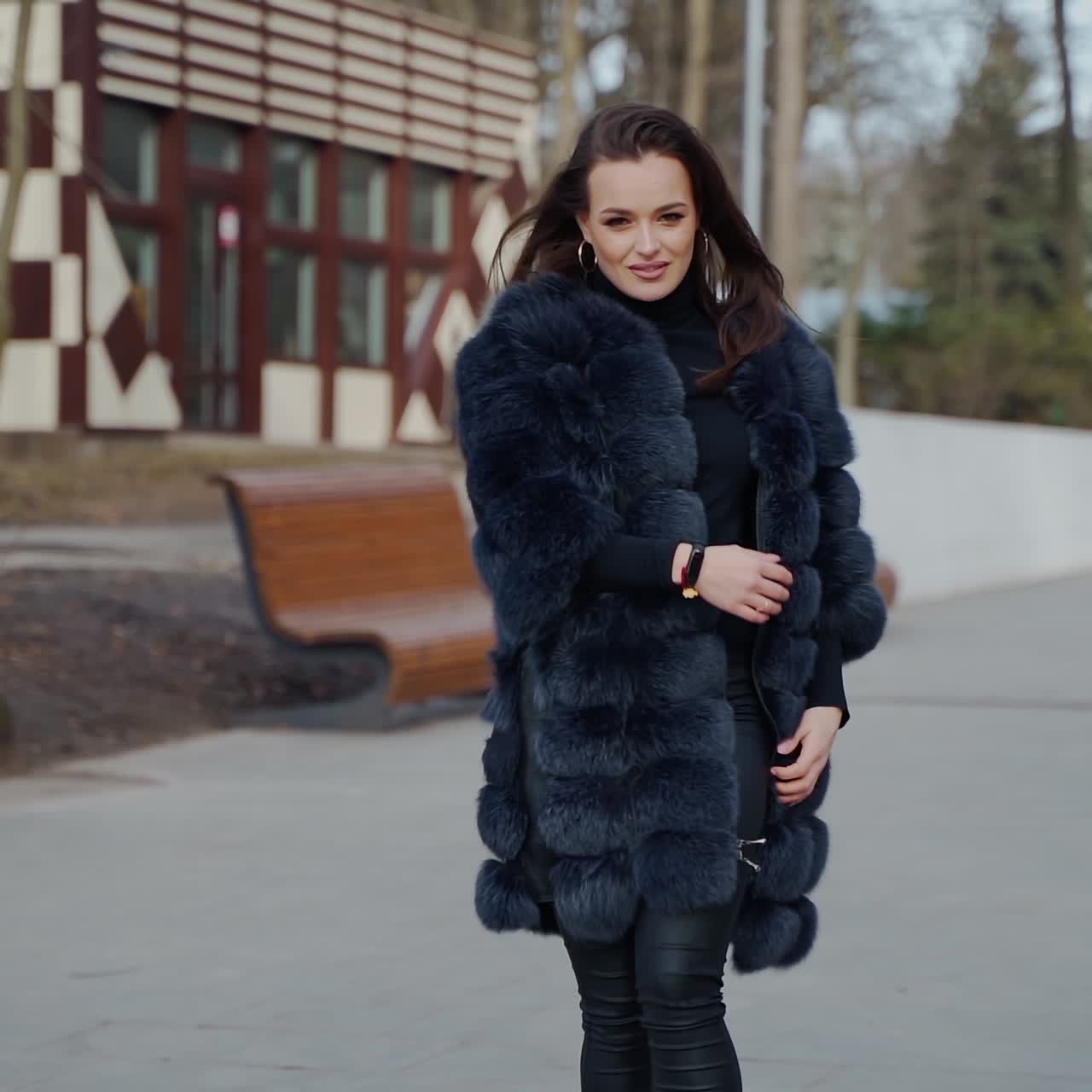 Attractive girl posing in the park. Beautiful young woman in black fur coat looking on camera and smiling outdoors. Slow motion.