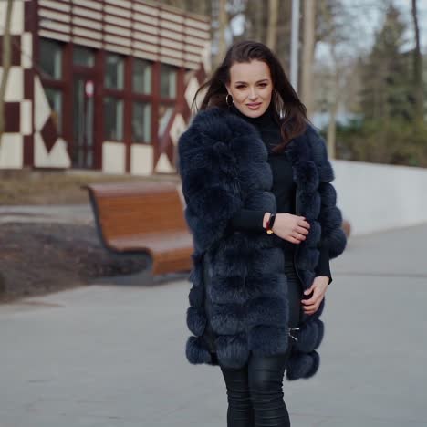 attractive girl posing in the park. beautiful young woman in black fur coat looking on camera and smiling outdoors. slow motion.