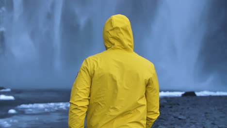 Young-Male-Traveler-in-yellow-rain-coat-in-front-of-huge-frozen-waterfall-in-slow-motion