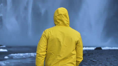 joven viajero con capa de lluvia amarilla frente a una enorme cascada congelada en cámara lenta