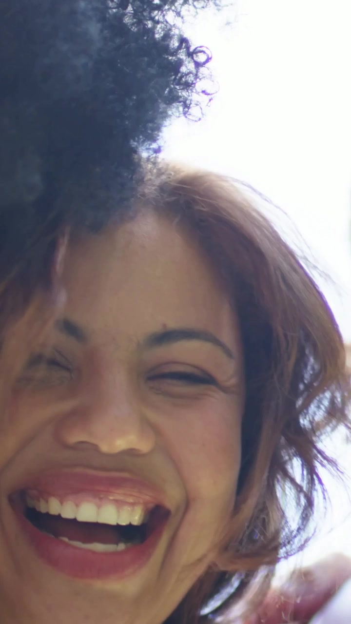 Video verticale: Due donne che ridono divertite, inclinate verso una telecamera bassa, all'aperto, una con maglietta bianca e l'altra con top rosso.