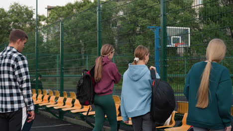 trainer waiting as students drop bags near fenced basketball court, one student turning toward coach, trees and court setting create athletic environment