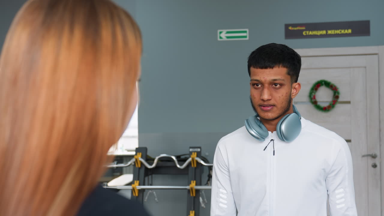 young woman nods agreement while communicating with friend inside gym as man walks away past weights and window city view, trainer with headphones around neck offers guidance under bright light