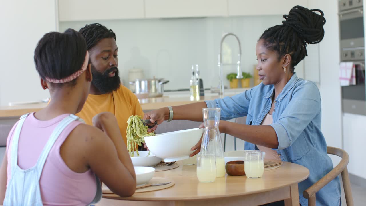African family mom scooping green pasta from white bowl for meal at dining table, father steadying