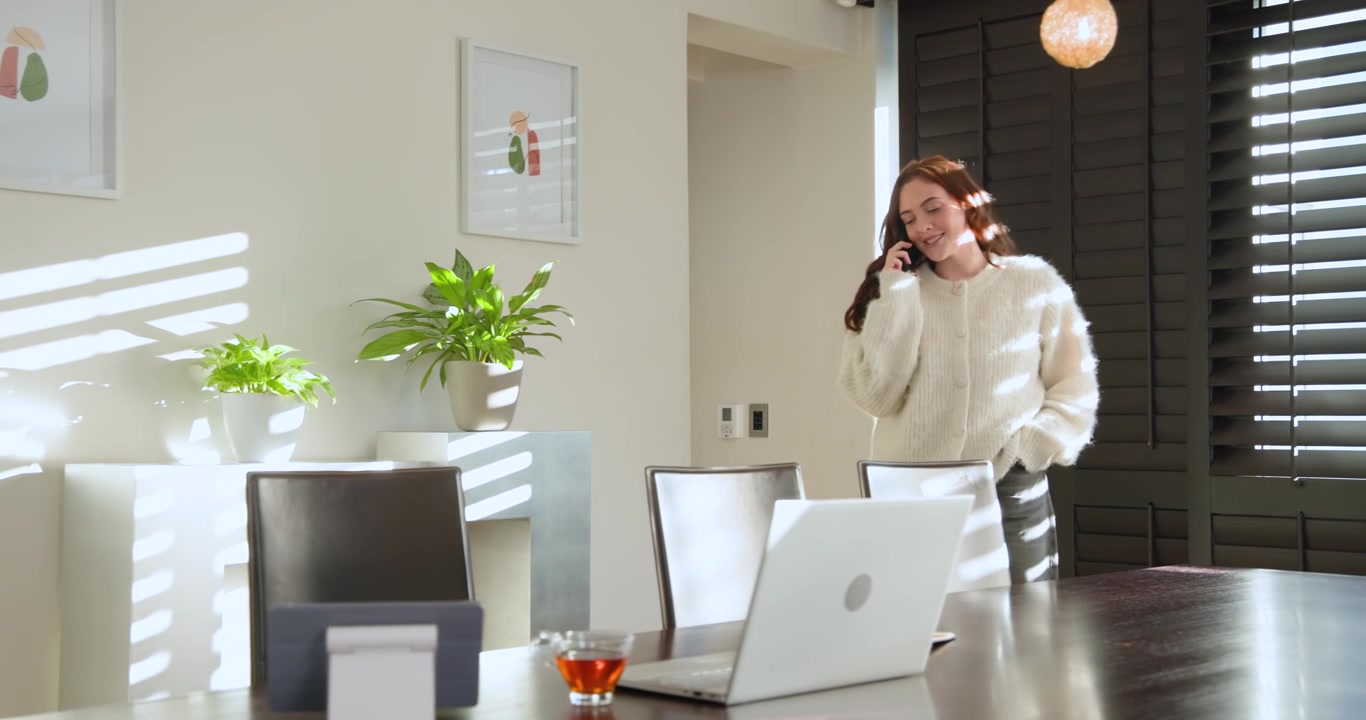 Donna che riceve una chiamata di lavoro al tavolo di casa, chinata, digita su un laptop argentato mentre controlla lo smartphone.