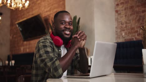 Black-person-folded-his-hands-on-the-table-in-front-of-a-laptop,-posing-and-looking-at-the-camera-while-in-a-cafe