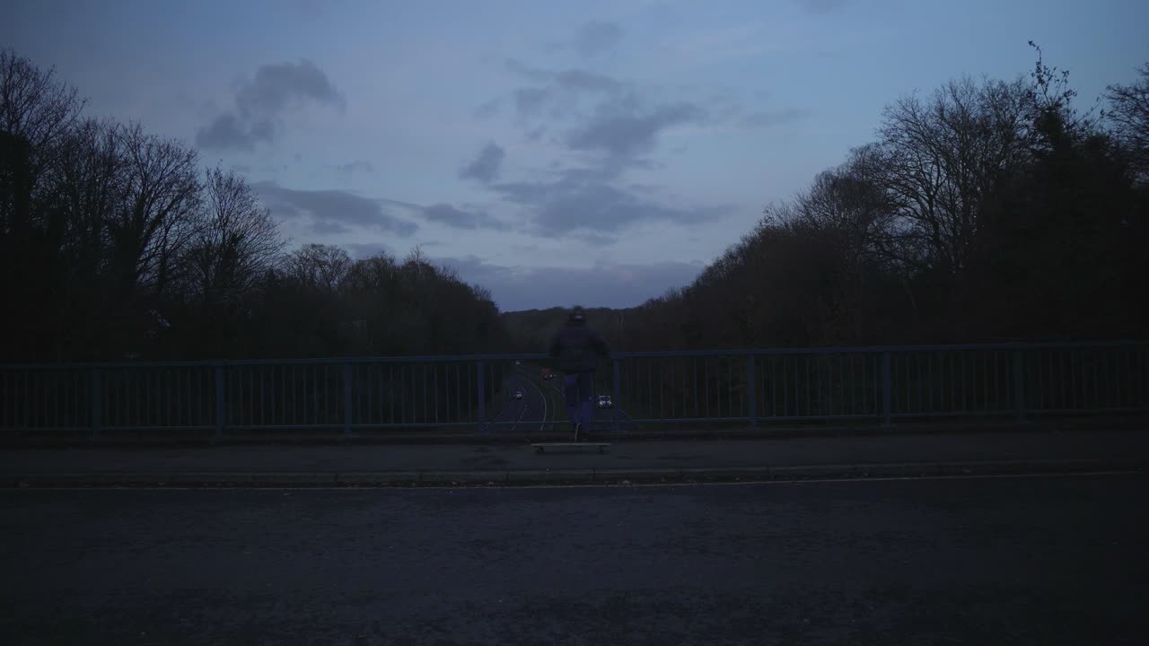 Skater leans up against guardrail in silhouette at dusk by river edge with ambient reflections, moody vibe