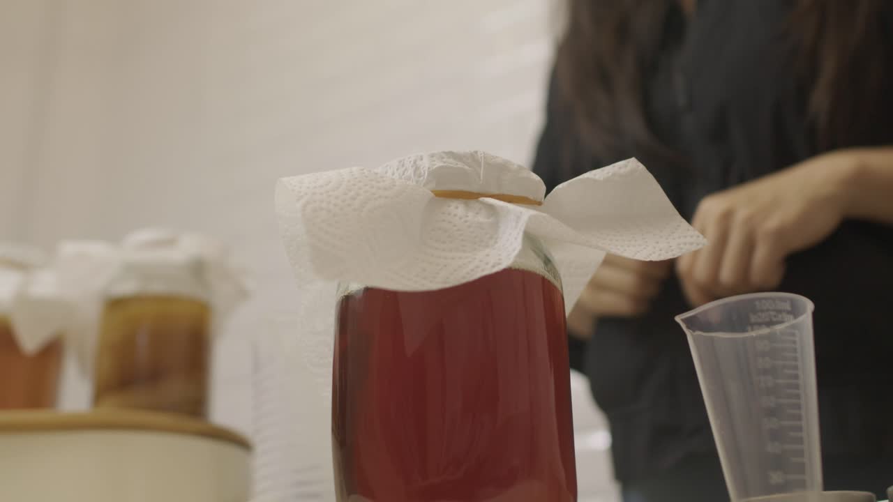 la mujer pone una cubierta de tejido en un frasco de vidrio con ingredientes de kombucha y la cierra con una banda de goma elástica