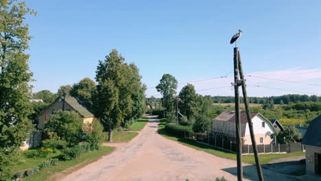 Una-Cigüeña-Se-Posa-En-Un-Poste-De-Electricidad-En-La-Tranquila-Plaza-De-Un-Pueblo-Báltico-Cerca-De-La-Frontera-Bielorrusa,-Captada-Por-Un-Dron