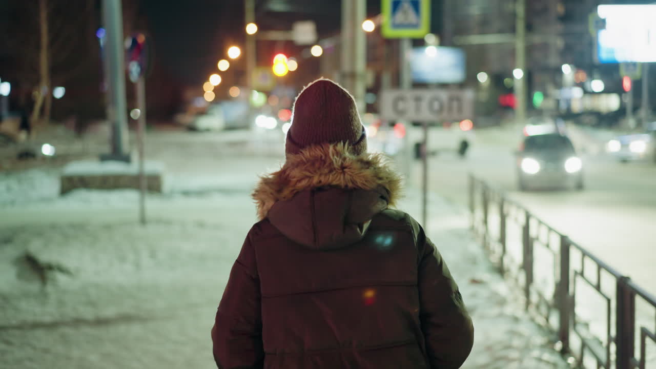 First-person view of a woman in a winter coat with a fur hood, walking at night in an urban setting. Blurry city lights and moving cars are visible in the background
