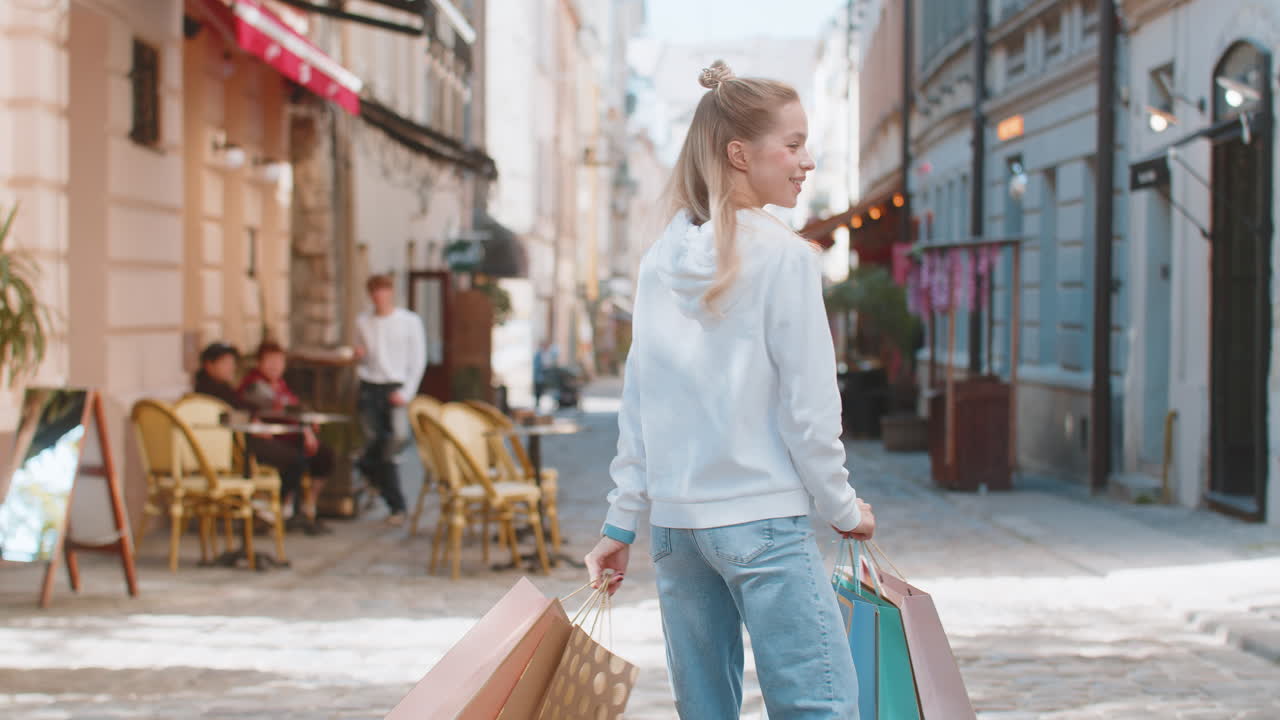 Happy smiling young caucasian woman shopaholic carrying shopping bags and walking on city street