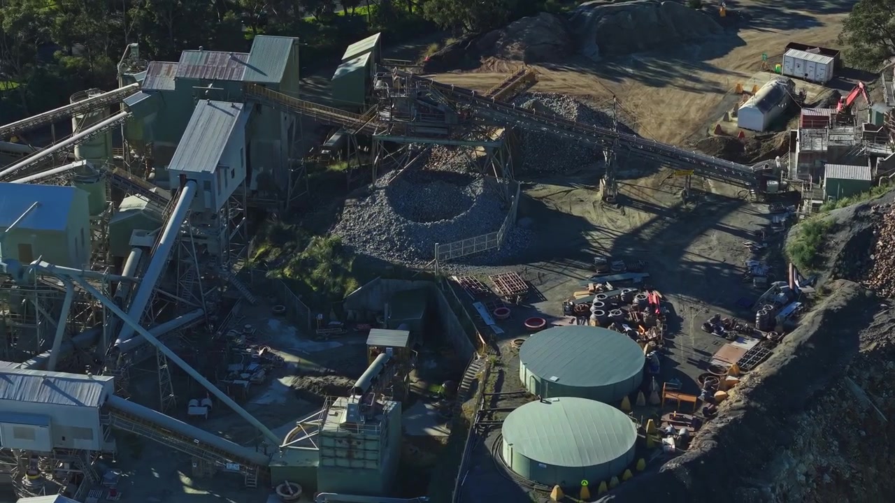 Dynamic aerial footage captures the expansive Montrose Quarry in Melbourne, featuring industrial structures, machinery, gravel piles, and circular tanks under bright daylight with clear shadows.