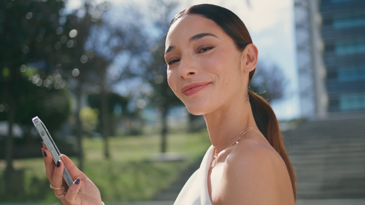 행복한 갈색 머리가 밖에서 소셜 미디어를 탐색하는 전화기를 가까이 보고 있습니다.