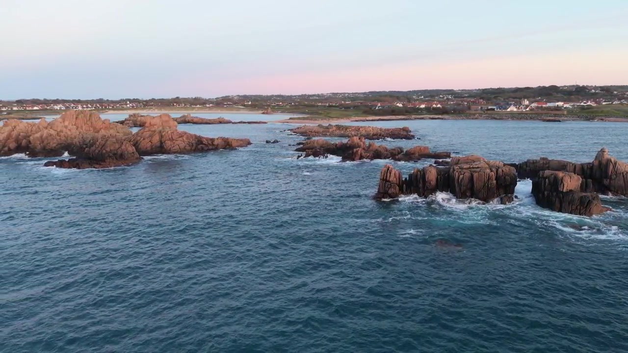 Guernsey, Isole del Canale, riprese panoramiche con drone di costa frastagliata con onde che si infrangono su affioramenti rocciosi di granito sotto il sole del tardo pomeriggio con il rosa che inizia a tingere l'orizzonte guardando verso la riva