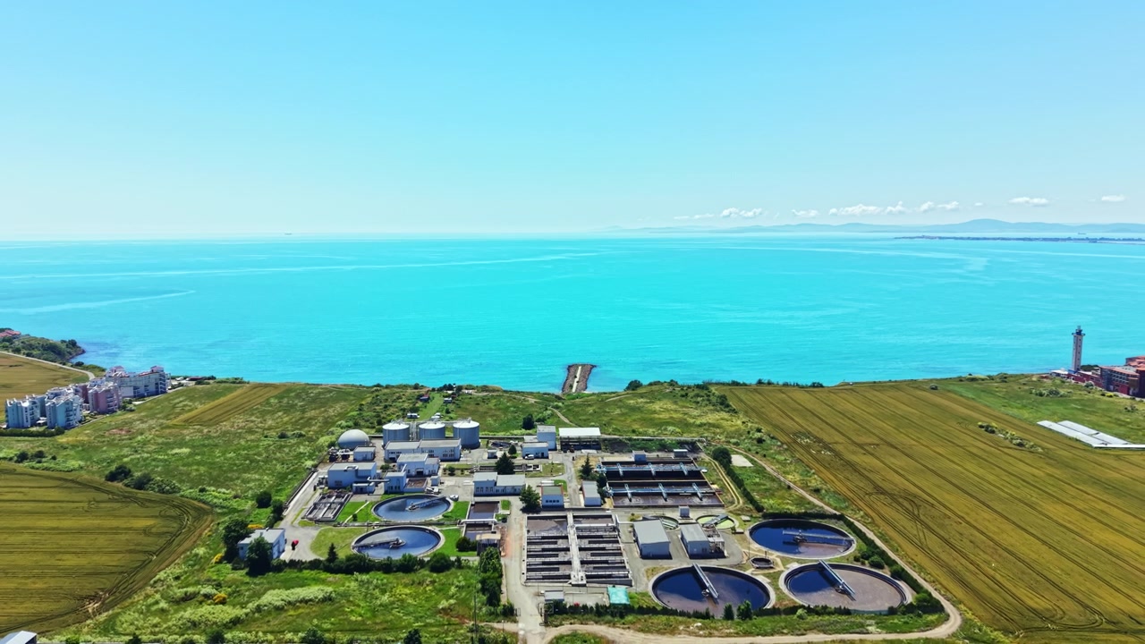 Water treatment plant near blue ocean under clear sky with green fields and buildings in the background at mid-afternoon