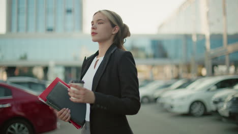 elegant young woman holding coffee cup and file folder walking through city parking lot with confident expression surrounded by modern buildings and parked cars in bright urban setting