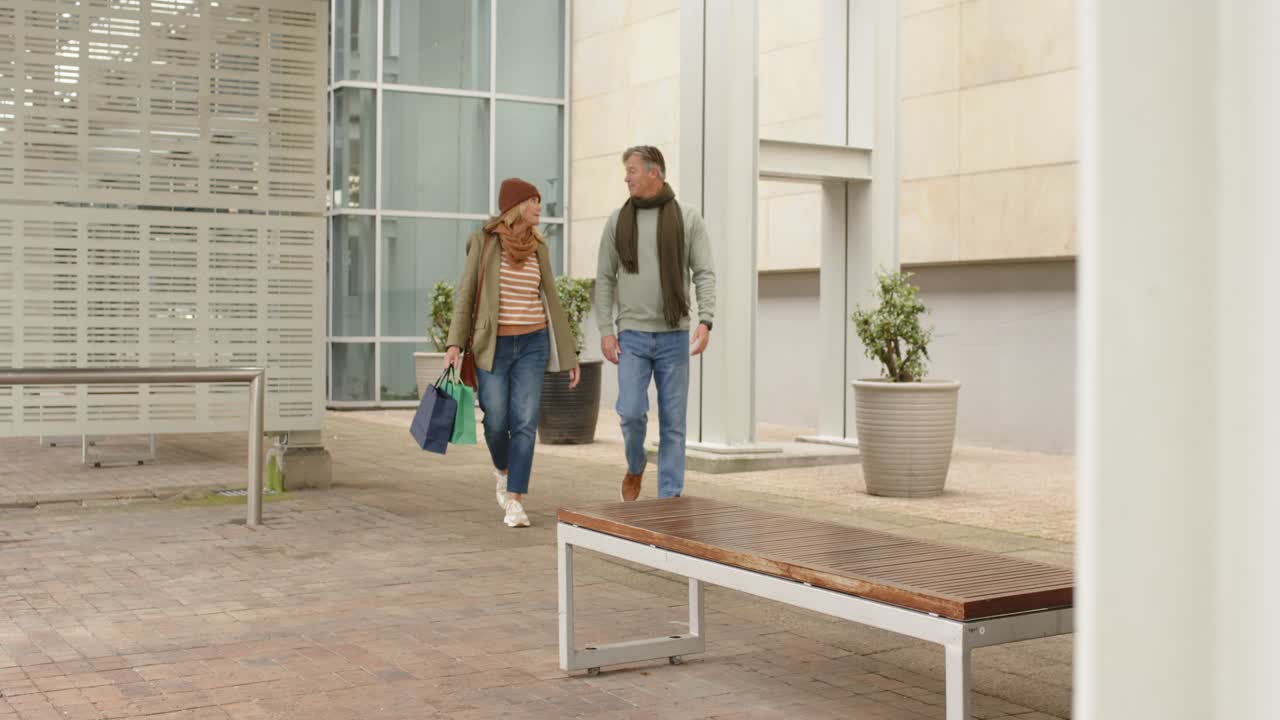 Coppia anziana che cammina in un cortile urbano dopo lo shopping, con borse, tenendo uno smartphone e conversando.