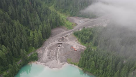 aerial view of a muddy landscape, the aftermath of a recent landslide, with construction equipment actively working on the site surrounded by dense alpine forests