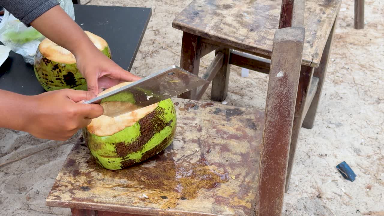 Close-up shot of hands using a large metal cleaver to chop open fresh green coconuts