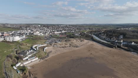 Wide aerial of Bude’s sandy beach, river mouth and Atlantic surf bordered by hillside homes, sports fields and holiday amenities, portraying vibrant seaside lifestyle and coastal recreation
