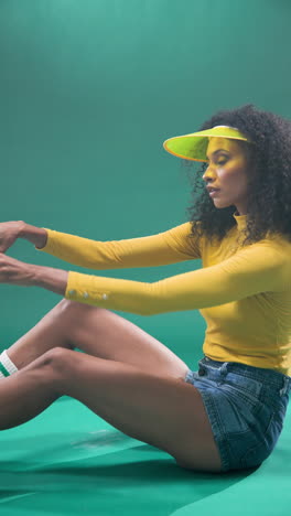 vertical video: woman in yellow top and visor holding sunglasses, sitting against green backdrop