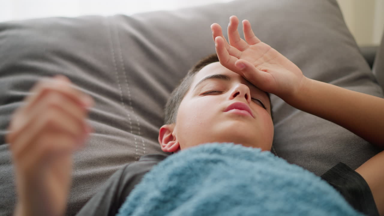 Close-up of sick child resting on bed, laying down with hand on head, looking unwell and tired, covered by blue blanket in soft, cozy indoor setting with bright window light