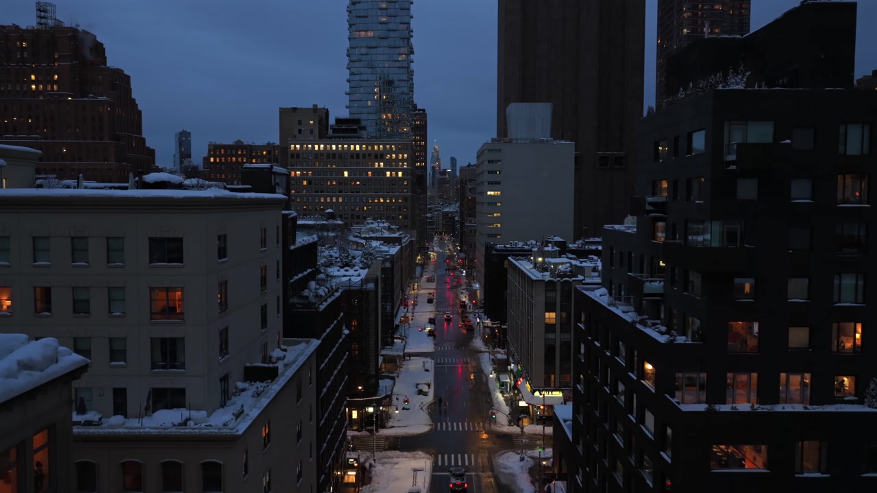 Aerial view rising above snowy residential street in downtown New York City at dusk, with glowing apartment windows, wet roads reflecting car lights and winter atmosphere between tall buildings.