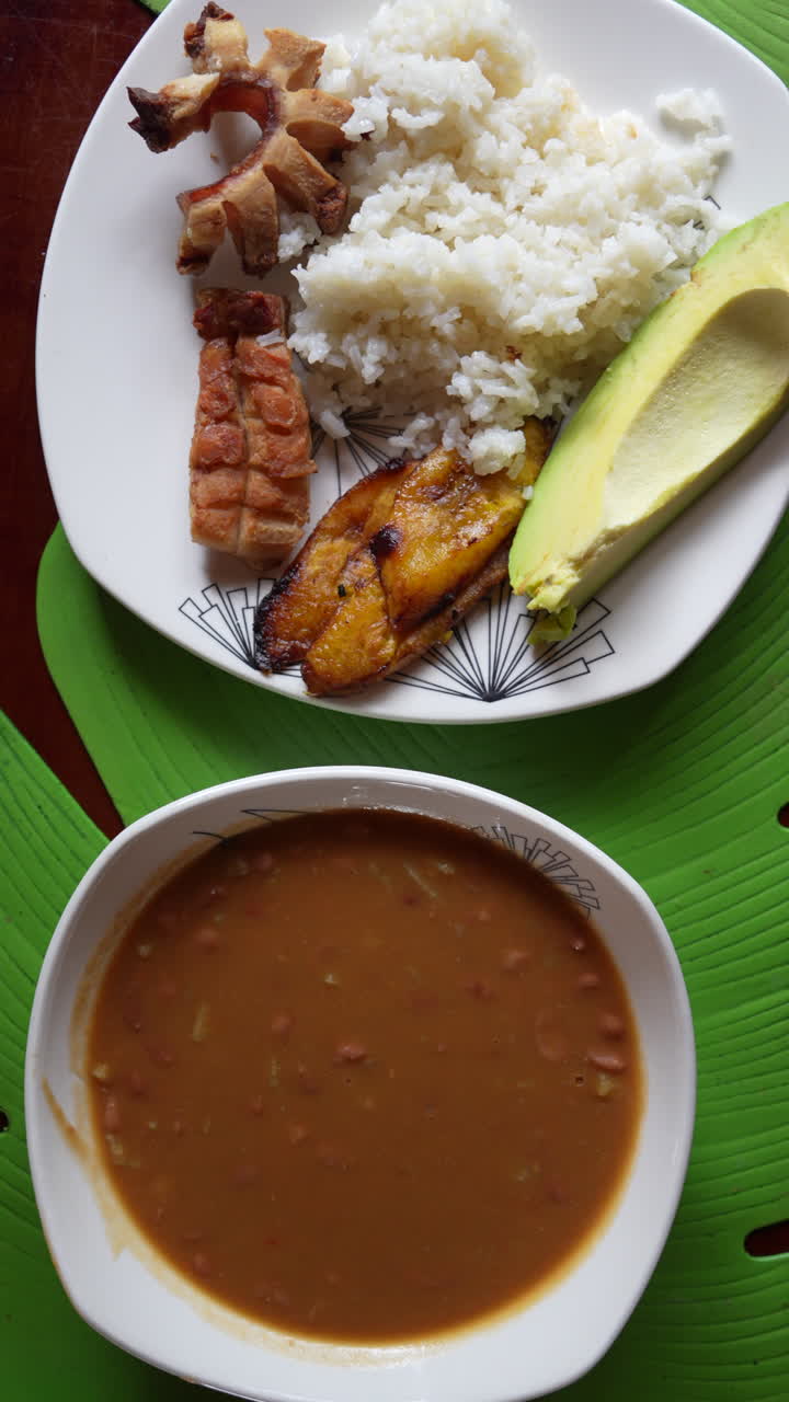 Vertical video showcasing a vibrant close-up shot of traditional Colombian bandeja paisa with savory beans, crispy pork, rice, plantain, and avocado. Perfect for mobile social media