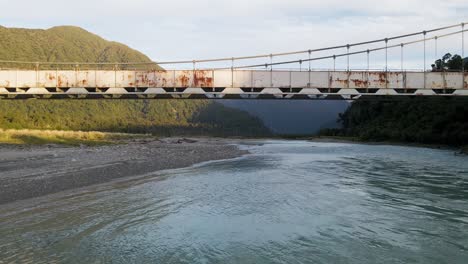 Aerial-dolly-out-revealing-steel-bridge-over-glacier-stream,-rugged-mountains-at-sunset