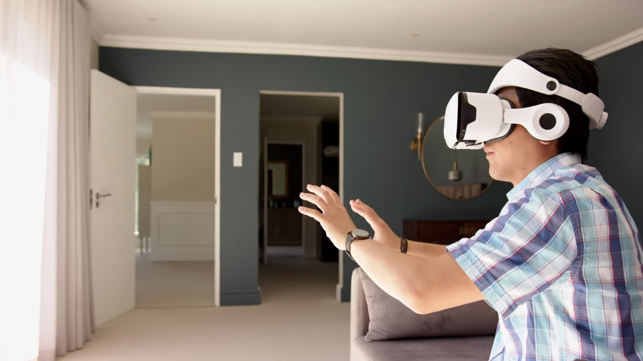 Using VR headset, asian man sitting on sofa and interacting with virtual environment, copy space