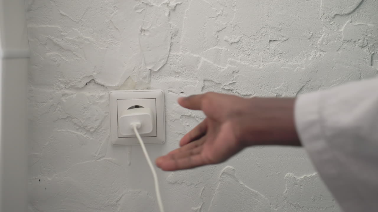 Youth in bathrobe reaching for charger plug, connecting device to outlet in modern bedroom. Soft lighting highlighting minimalist decor and calm atmosphere, morning routine setting