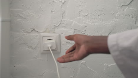 youth in bathrobe reaching for charger plug, connecting device to outlet in modern bedroom. soft lighting highlighting minimalist decor and calm atmosphere, morning routine setting
