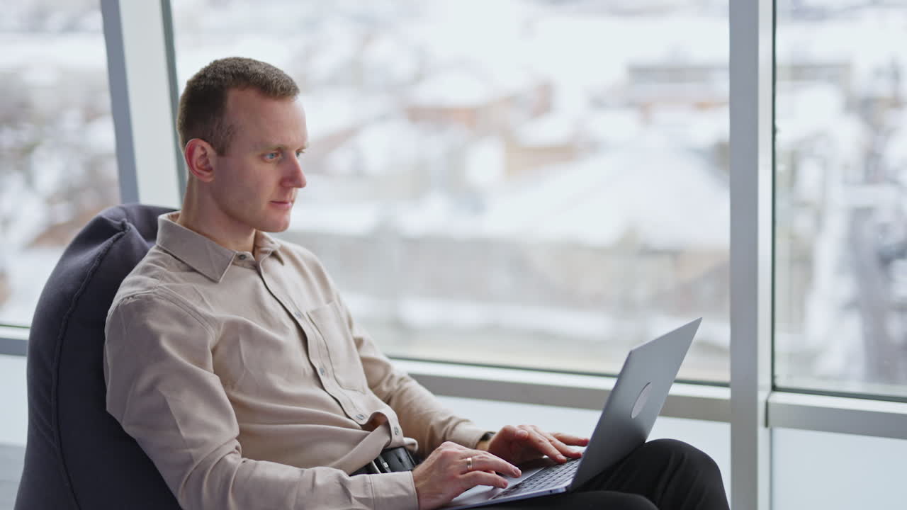 Caucasian male sitting in armchair and holding a laptop on his knees. Working man at the backdrop of panoramic window.