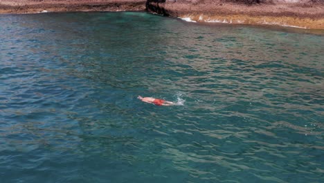 Man-snorkeling-in-blue-tropical-water-at-rocky-cliff-shore,-aerial